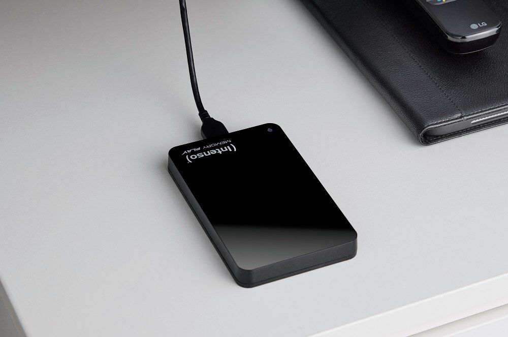 Na białym biurku leży czarny dysk zewnętrzny z napisem \'Intenso Memory Play\', podłączony kablem USB. Obok znajduje się czarne etui na tablet.