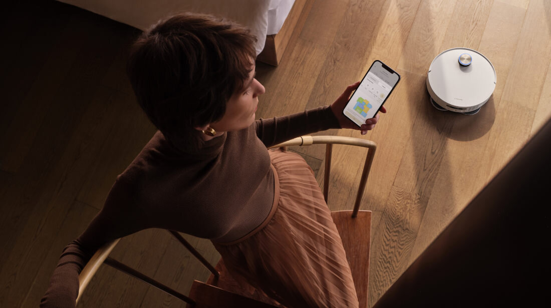 Osoba siedzi na krześle, trzymając w ręku smartfon z wyświetloną aplikacją. Na podłodze widać biały robot sprzątający.