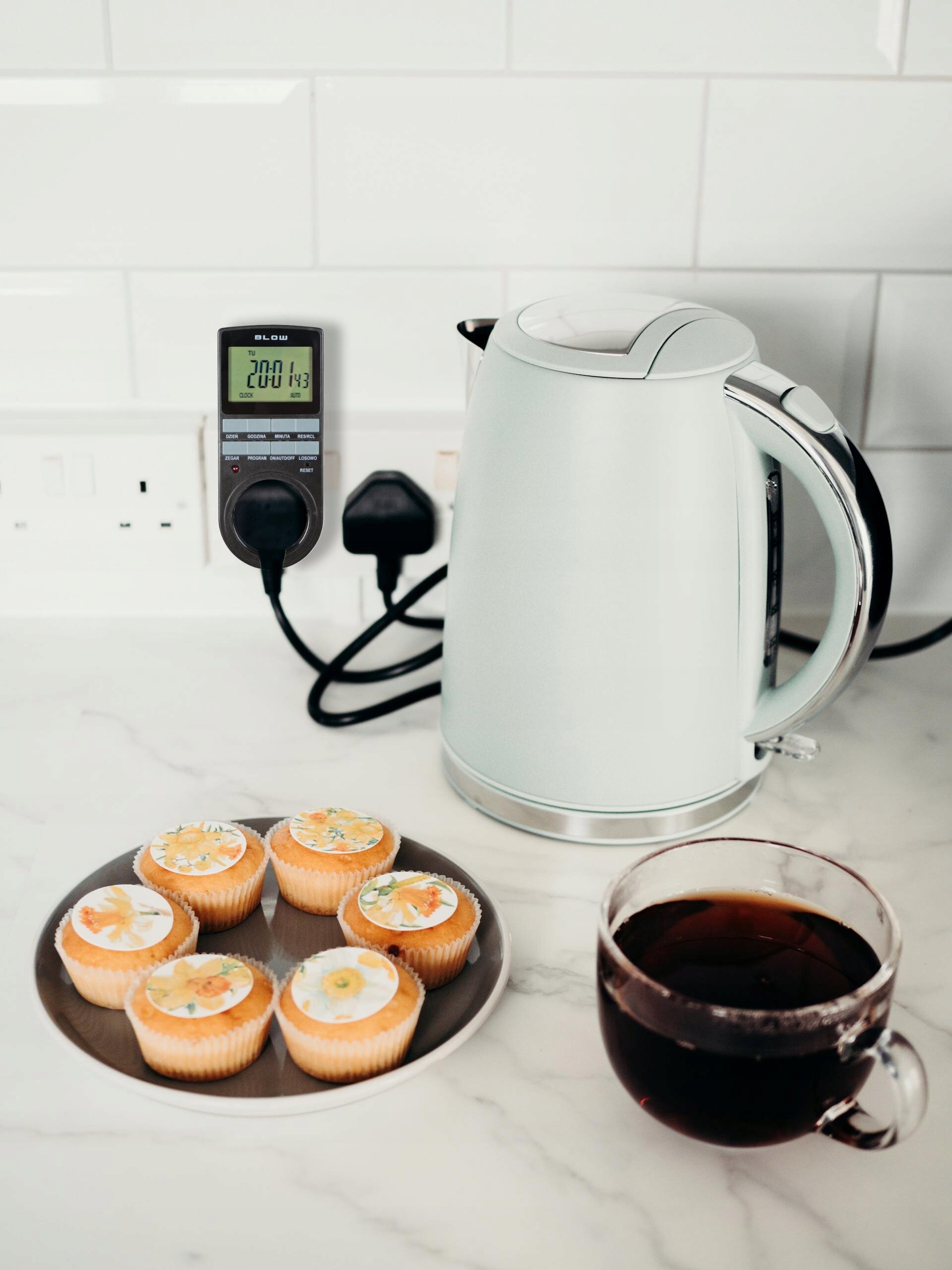 Na blacie kuchennym stoi czajnik elektryczny obok programatora czasowego z wyświetlaczem LCD, na którym widnieje napis \'BLOW\' oraz liczby \'20:01 143\'. Obok znajduje się talerz z sześcioma babeczkami i filiżanka z ciemnym napojem.