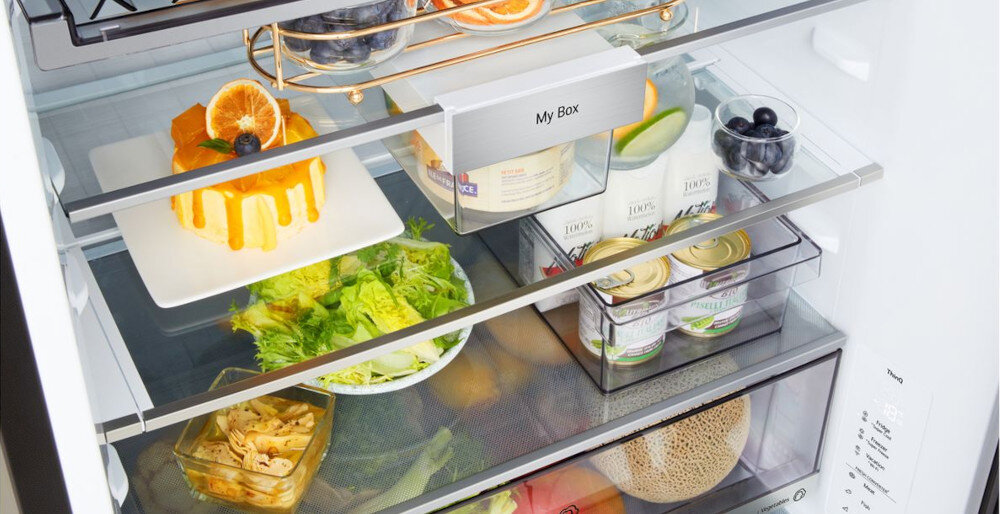 Wnętrze lodówki z trzema półkami, na których znajdują się różne produkty spożywcze, w tym ciasto, sałata, owoce i puszki. Na jednej z półek widoczny jest napis \'My Box\'.