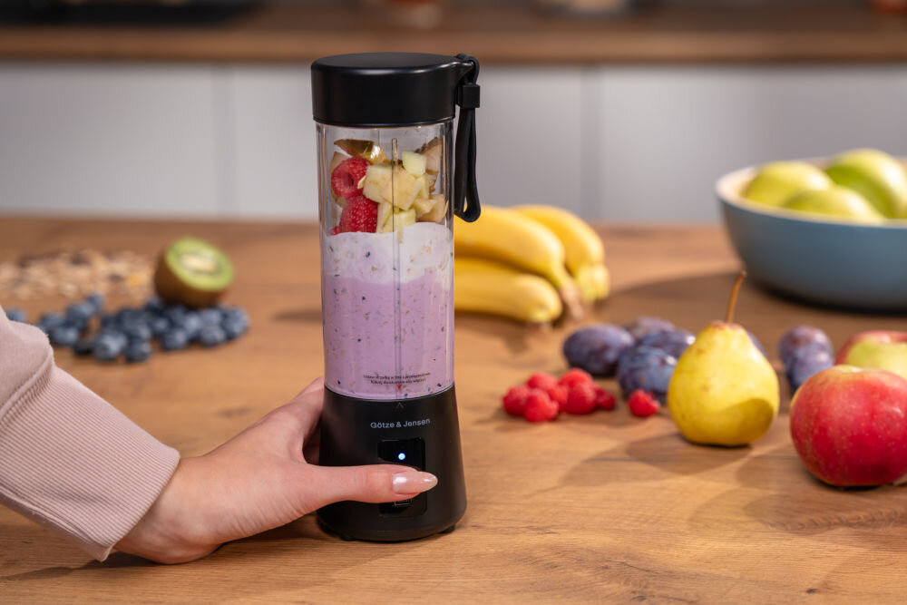 Na drewnianym blacie stoi czarny blender personalny z logo \'Götze & Jensen\', wypełniony owocami i jogurtem. W tle widoczne są różne owoce, takie jak banany, jabłka i kiwi.
