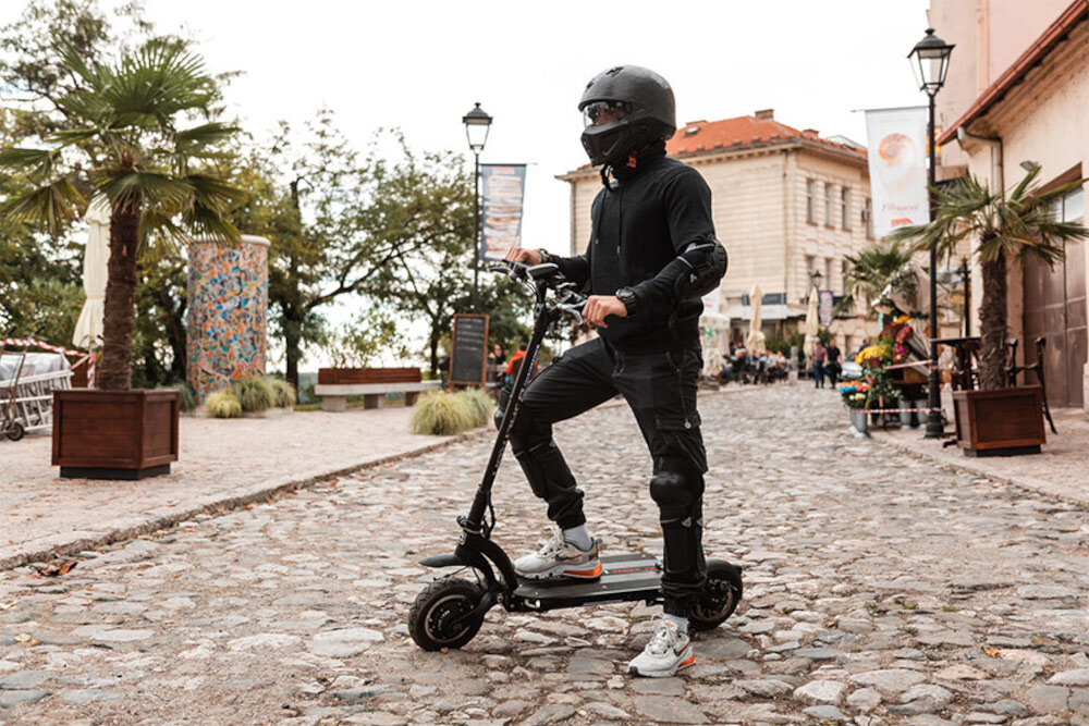 Osoba w kasku stoi na czarnej hulajnodze elektrycznej na brukowanej ulicy, otoczonej roślinnością i budynkami. W tle widoczne są palmy i kolorowe dekoracje.
