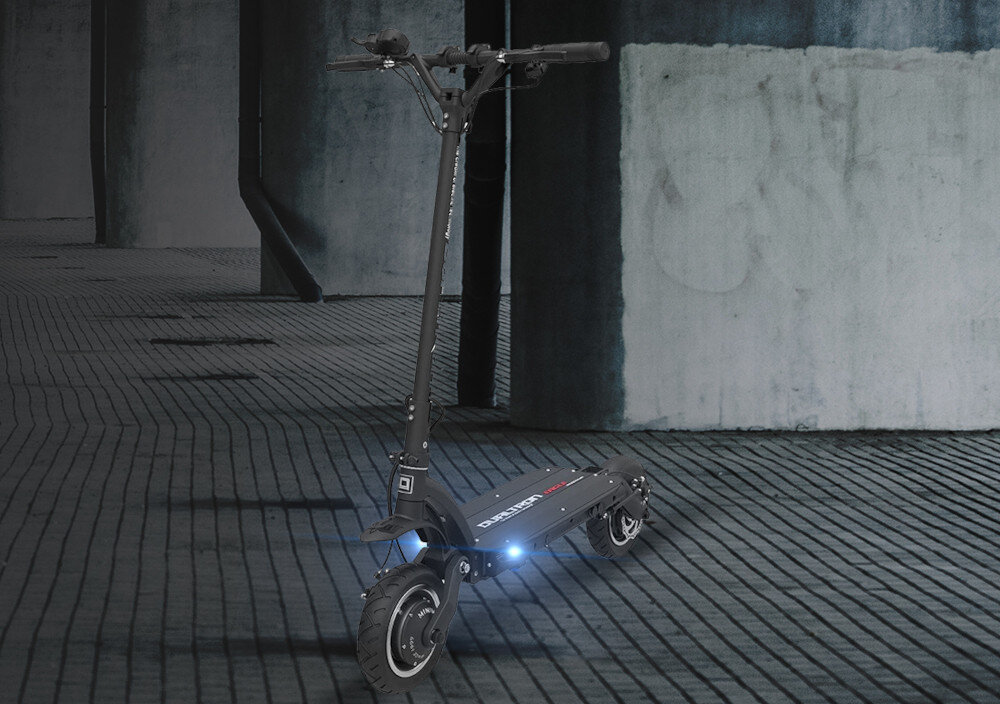 Czarna hulajnoga elektryczna stoi na tle betonowej ściany, z widocznym logo \'DUALTRON\' na platformie. Przednie światło hulajnogi jest włączone.