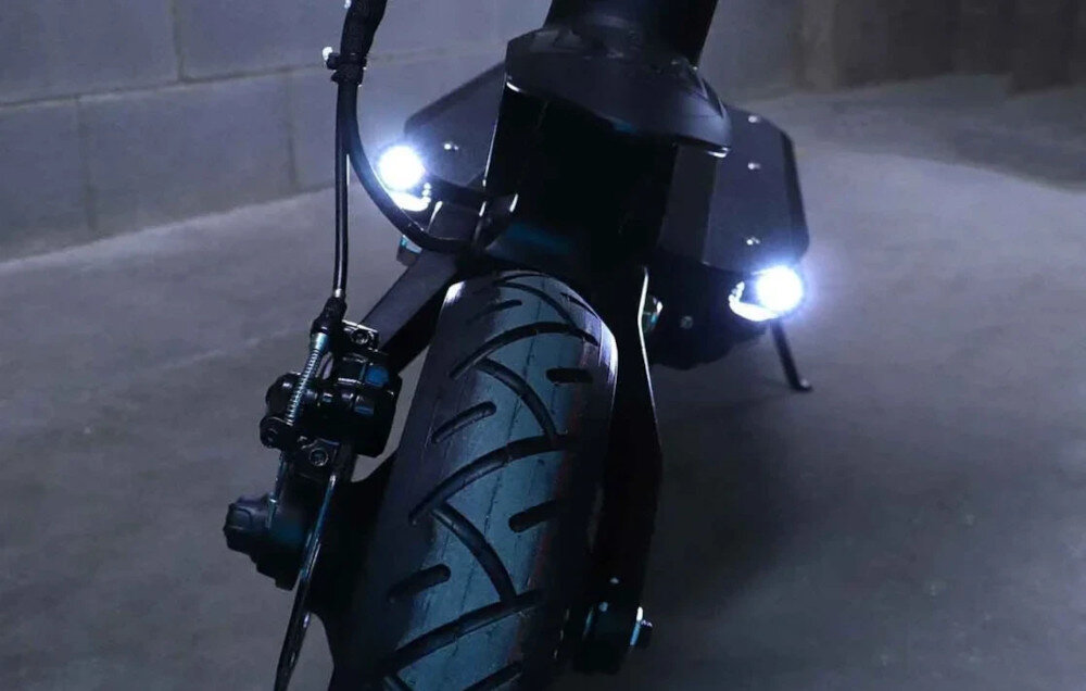 Czarna hulajnoga elektryczna z widocznym przednim kołem i dwoma świecącymi reflektorami. W tle znajduje się szara ściana.