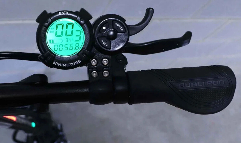 Na zdjęciu widoczna jest kierownica hulajnogi elektrycznej z wyświetlaczem pokazującym wartości \'003\', \'34°\', \'0056.8\'. Obok wyświetlacza znajdują się przyciski z napisami \'POWER\' i \'MODE\', a na uchwycie widnieje napis \'DUALTRON\'.