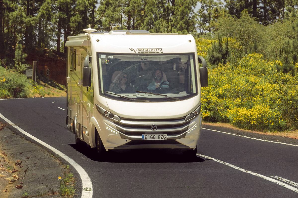 Na zdjęciu widać biały kamper marki Mobilvetta jadący po asfaltowej drodze otoczonej zielenią i żółtymi kwiatami. Na przodzie pojazdu widoczna jest tablica rejestracyjna z numerem 6168 KZG.