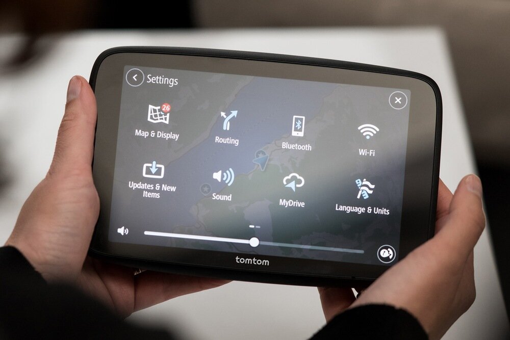 Osoba trzyma urządzenie nawigacyjne z wyświetlonym menu ustawień, zawierającym ikony takie jak Map & Display, Routing, Bluetooth, Wi-Fi, Updates & New Items, Sound, MyDrive oraz Language & Units. Na dole ekranu widoczny jest pasek regulacji głośności, a na obudowie znajduje się logo \'tomtom\'.