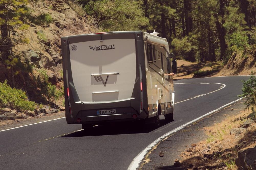 Kamper marki Mobilvetta jedzie po krętej, asfaltowej drodze otoczonej drzewami i skałami. Na tylnej części pojazdu widoczna jest tablica rejestracyjna z numerem 6168 KZG.