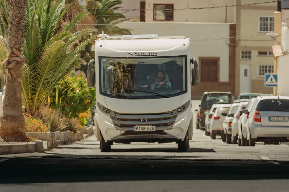 Duży biały kamper z napisem \'Mobilvetta\' na przodzie jedzie ulicą otoczoną palmami i zaparkowanymi samochodami. Na tablicy rejestracyjnej widnieje numer 6168 KZG.