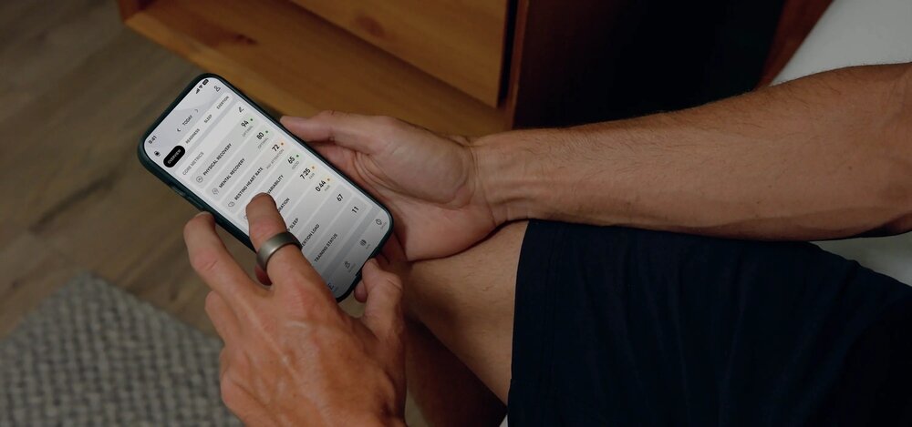 Osoba trzyma smartfon z wyświetlonym interfejsem aplikacji, na palcu ma założony pierścień. Na ekranie widoczne są różne dane liczbowe i tekstowe.
