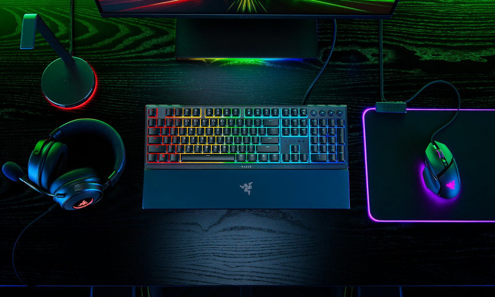 Na ciemnym biurku znajduje się klawiatura z podświetleniem RGB, mysz na podkładce oraz słuchawki. Logo Razer widoczne jest na klawiaturze i podkładce.
