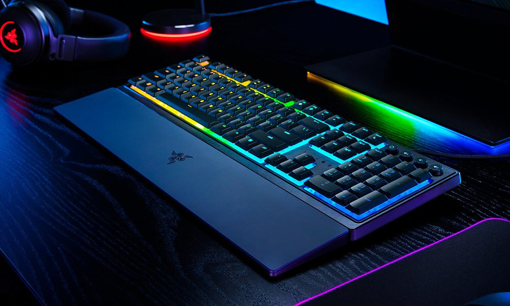 Klawiatura z podświetleniem RGB, z widocznym logo RAZER, umieszczona na ciemnym biurku obok słuchawek i laptopa.