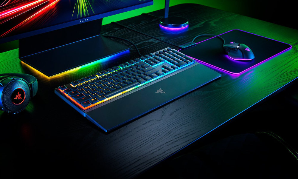 Na biurku znajduje się klawiatura z podświetleniem RGB, mysz oraz monitor. Logo RAZER widoczne jest na klawiaturze i myszce.