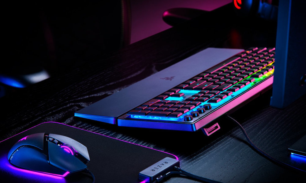 Na biurku znajduje się klawiatura z podświetleniem RGB oraz myszka na podkładce. Na klawiaturze widoczne jest logo producenta.