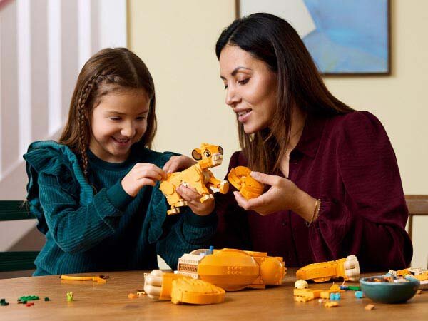 Dwie osoby składają zestaw klocków LEGO przedstawiający młodego Simbę. Na stole znajdują się elementy zestawu w kolorze żółtym i zielonym.