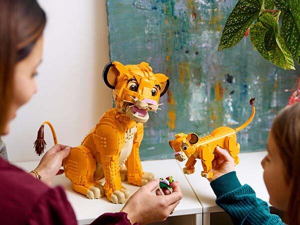 Na obrazie widać dwie figurki LEGO przedstawiające młodego Simbę z filmu Król Lew. Figurka jest w kolorze pomarańczowym z czarnymi i białymi detalami, a w tle znajduje się kolorowy obraz na ścianie.