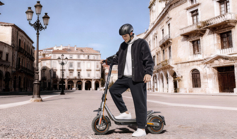 Osoba w kasku stoi obok czarnej hulajnogi elektrycznej na tle zabytkowych budynków. Hulajnoga ma duże koła i pomarańczowe akcenty na oponach.