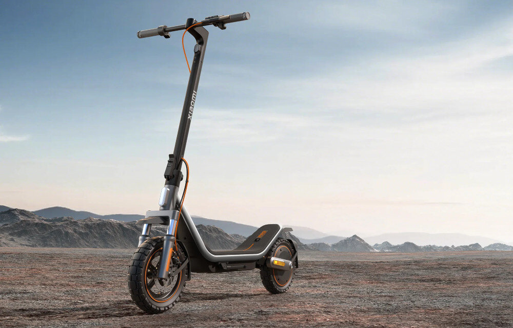 Czarna hulajnoga elektryczna z pomarańczowymi akcentami stoi na pustynnym terenie. Na kierownicy widoczny jest napis \'XIAOMI\'.