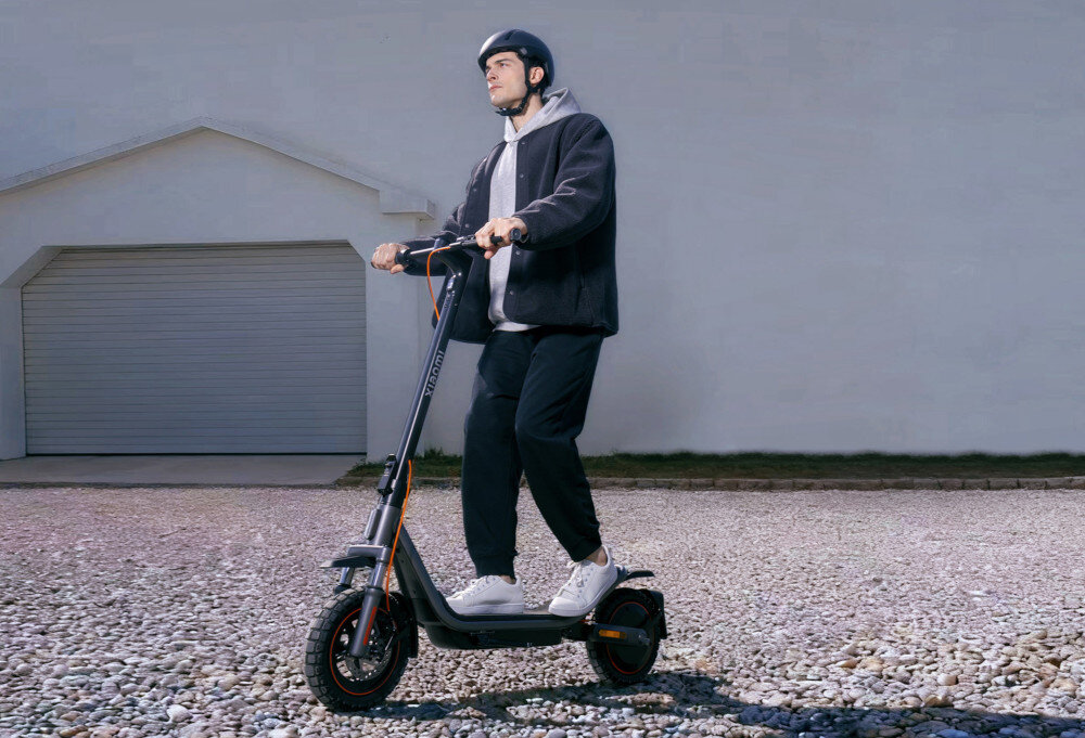 Osoba w kasku jedzie na czarnej hulajnodze elektrycznej po żwirowej nawierzchni. W tle widoczna jest biała ściana budynku z zamkniętą bramą garażową.