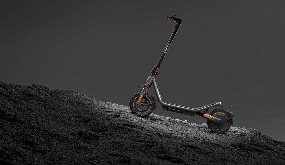 Czarna hulajnoga elektryczna z pomarańczowymi akcentami stoi na skalistym terenie. Na kierownicy widoczny jest napis \'XIAOMI\'.