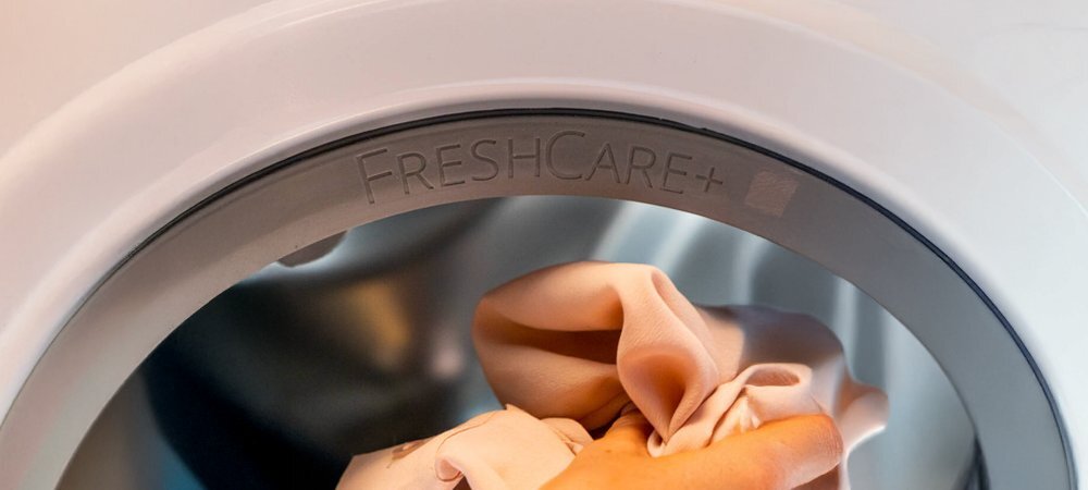 Zbliżenie na drzwiczki pralki z widocznym napisem \'FRESHCARE+\'. Wewnątrz bęben pralki, do którego wkładane są jasne tkaniny.