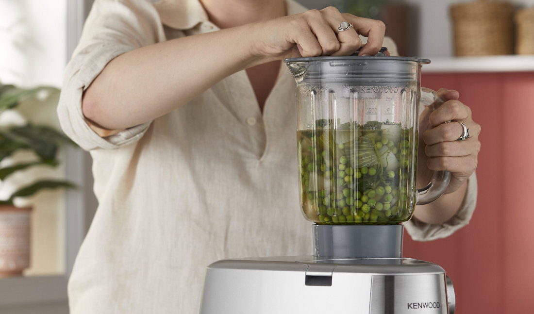 Osoba obsługuje blender kielichowy z zielonymi warzywami, umieszczony na robocie kuchennym marki KENWOOD. W tle widoczne są elementy kuchenne, takie jak rośliny i kosze.