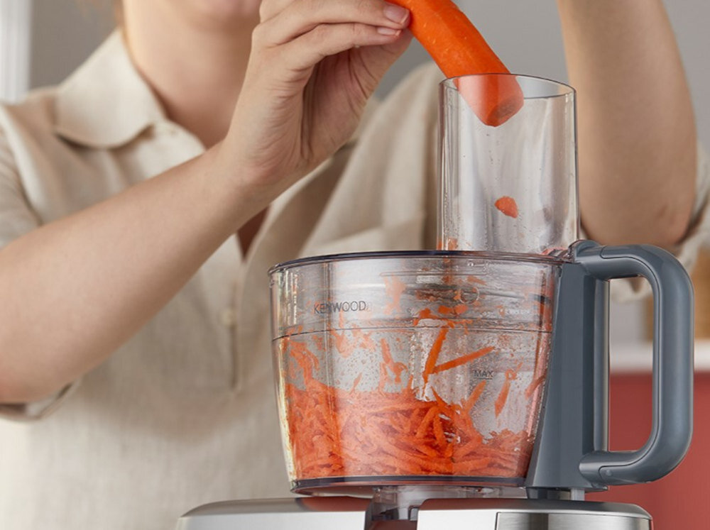 Osoba wkłada marchewkę do robota kuchennego marki Kenwood, w którym znajdują się już starte kawałki marchewki. Na pojemniku widoczne jest logo \'KENWOOD\'.