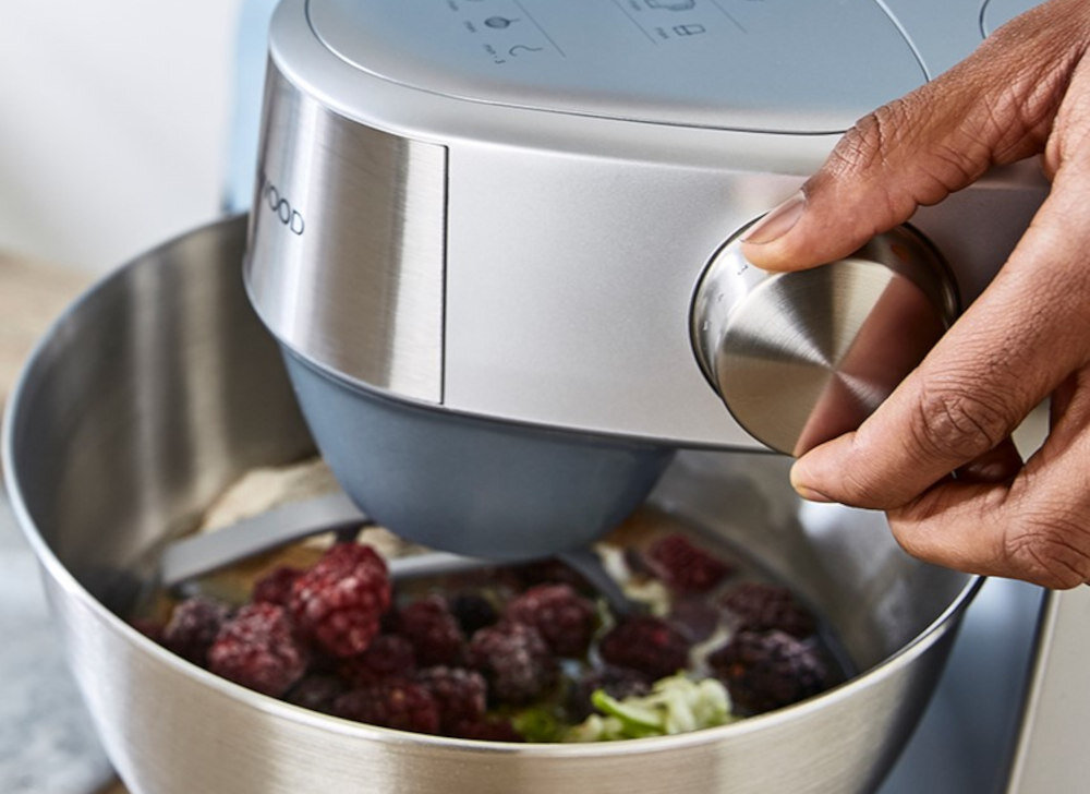 Osoba obsługuje robot kuchenny z metalową misą, w której znajdują się owoce i inne składniki. Na urządzeniu widoczne jest logo \'KENWOOD\'.