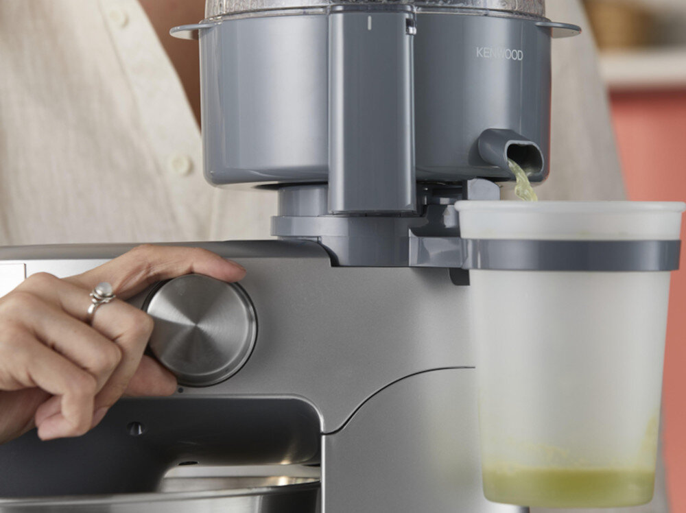 Osoba obsługuje robot kuchenny, obracając pokrętło. Na urządzeniu widoczne jest logo \'KENWOOD\', a do pojemnika spływa zielony płyn.