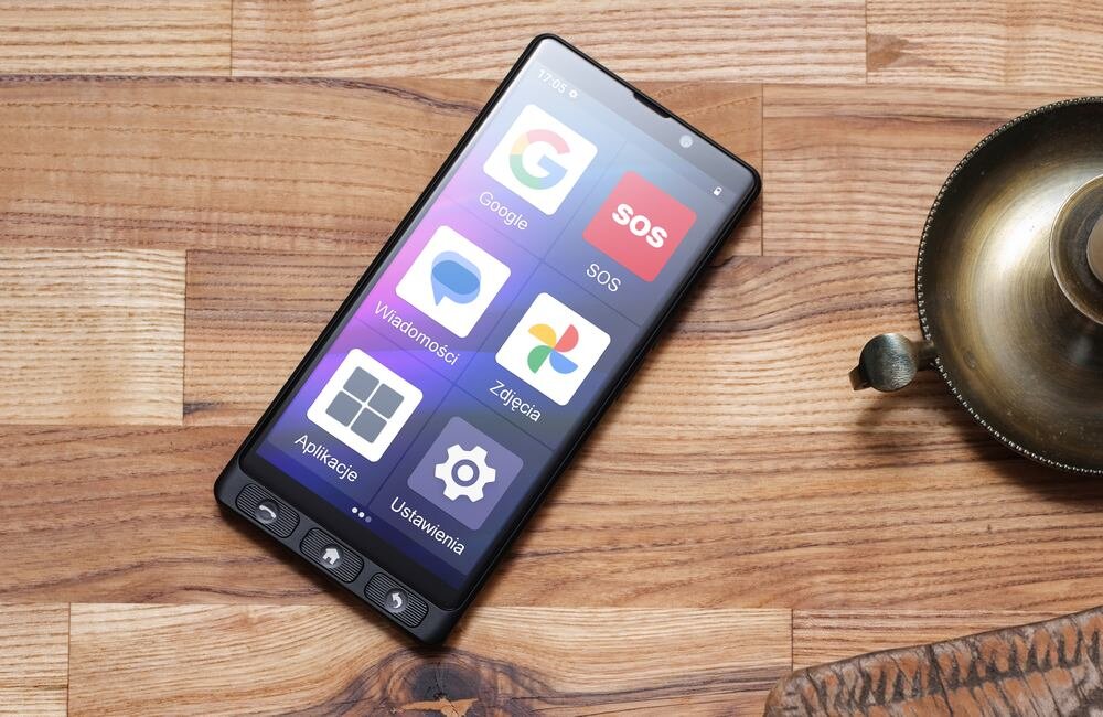 Smartfon leży na drewnianej powierzchni, na ekranie widoczne są ikony aplikacji: Google, SOS, Wiadomości, Zdjęcia, Aplikacje, Ustawienia. Obok smartfona znajduje się metalowy przedmiot przypominający lampę.