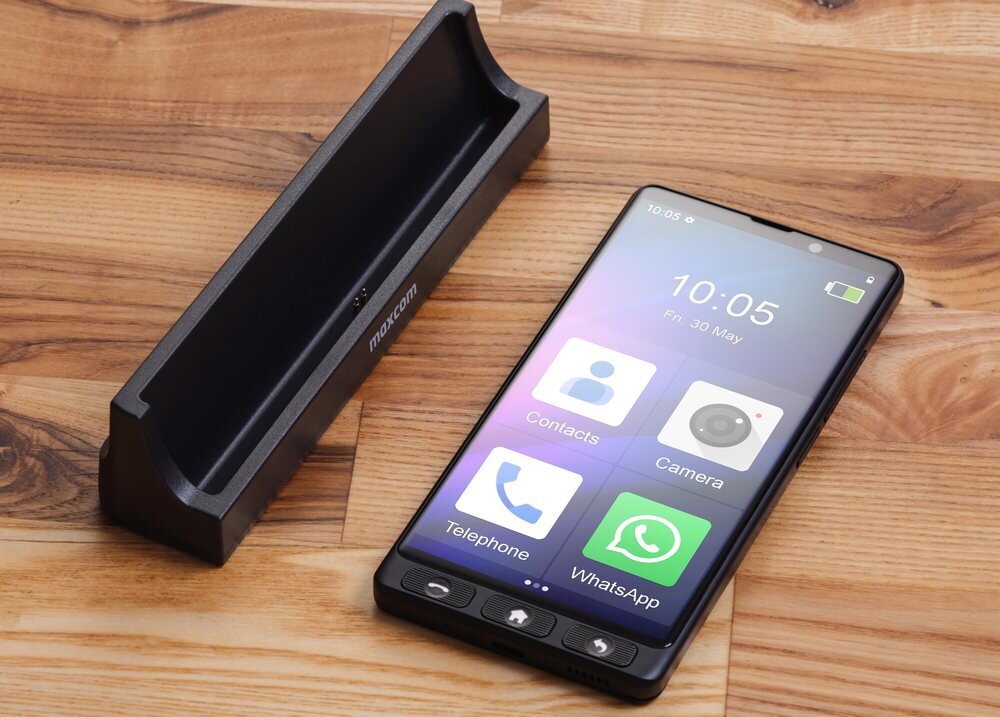 Na drewnianym tle leży smartfon z wyświetlaczem pokazującym ikonki aplikacji takich jak Contacts, Camera, Telephone i WhatsApp oraz godzinę 10:05, piątek, 30 maja. Obok znajduje się czarna podstawka z napisem \'maxcom\'.
