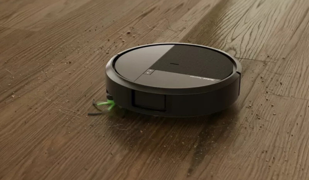 Czarny robot sprzątający porusza się po drewnianej podłodze, zbierając drobne zanieczyszczenia. Widać zielone szczotki wystające spod urządzenia.