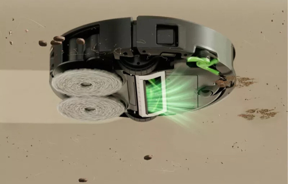Obraz przedstawia spód czarnego robota sprzątającego z dwoma okrągłymi szczotkami i zielonym światłem emitowanym z otworu. Wokół robota unoszą się drobinki kurzu i brudu.