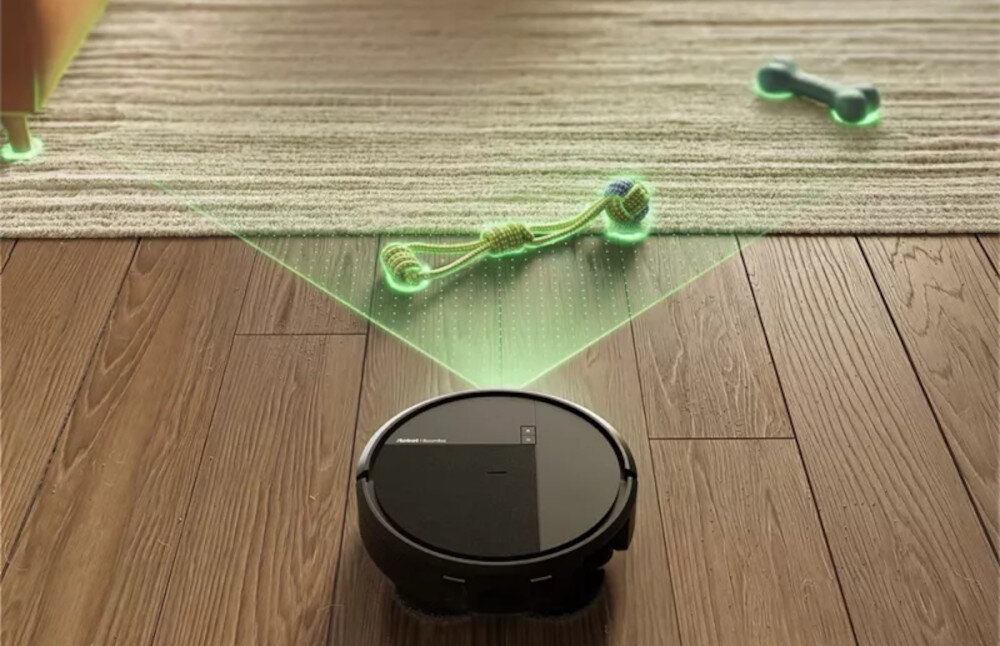 Czarny robot sprzątający porusza się po drewnianej podłodze, zbliżając się do dywanu, na którym leżą dwie zabawki. Na robocie widoczne jest logo \'Roomba Combo\'.