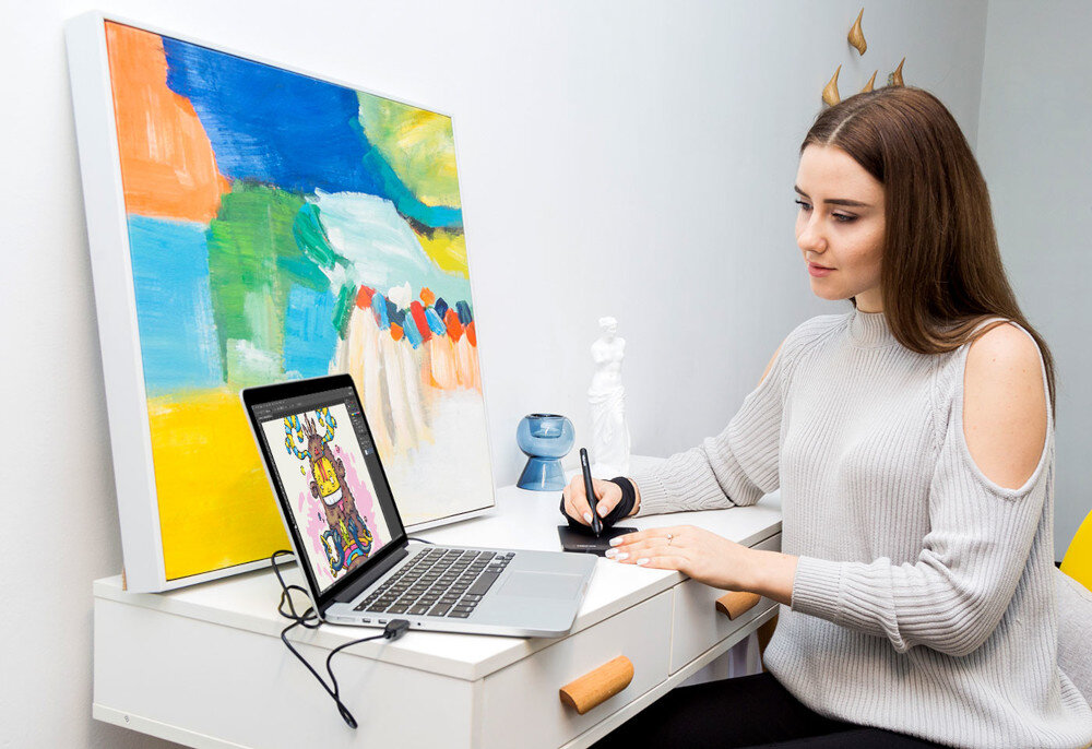 Osoba siedzi przy biurku, używając tabletu graficznego podłączonego do laptopa, na którym wyświetlany jest kolorowy rysunek. Na biurku znajduje się również kolorowy obraz oraz mała rzeźba.