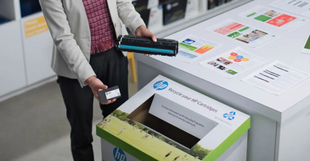 Osoba trzyma toner i telefon nad pojemnikiem z napisem \'Recycle your HP Cartridges\'. W tle widoczne są ulotki i broszury na białym blacie.