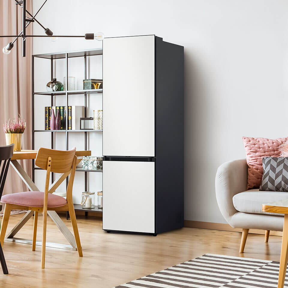 Przedstawiony na zdjęciu model Samsung Bespoke RB38C7B6BAP należy do linii lodówek o wysokości 2030 mm Wnętrze nowoczesnego salonu z lodówką o prostym, eleganckim designie w kolorze białym i czarnym. Obok lodówki znajduje się regał z książkami i dekoracjami, a w pomieszczeniu widoczne są także stół, krzesła i sofa z poduszkami.