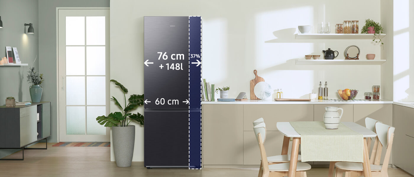 W kuchni znajduje się lodówka w kolorze grafitowej stali z oznaczeniami wymiarów: 76 cm + 148 l, 60 cm, 37%. Obok lodówki widoczny jest stół z krzesłami oraz półki z naczyniami i dekoracjami.