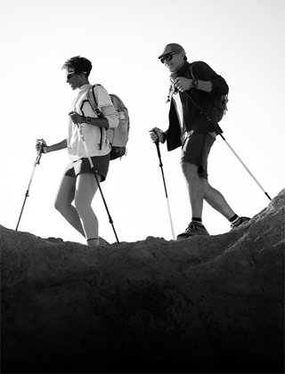 Czarno-białe zdjęcie przedstawia dwie osoby wędrujące po skalistym terenie z kijkami trekkingowymi. Obie osoby mają na sobie plecaki i sportowe ubrania.