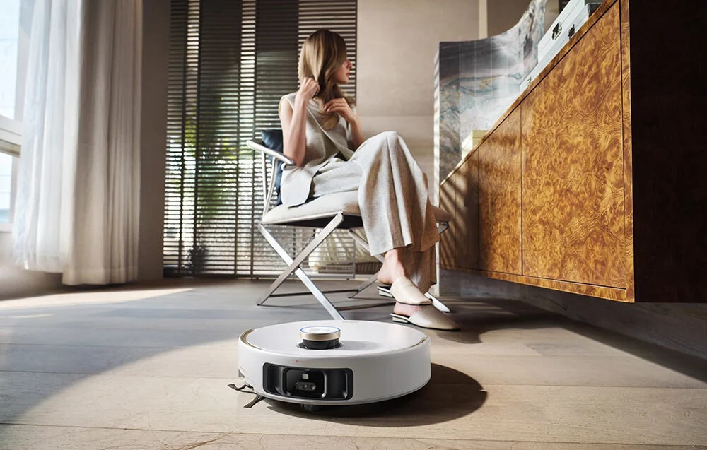 Na podłodze w jasnym pomieszczeniu znajduje się biały robot sprzątający. W tle siedzi osoba na krześle przy drewnianej szafce.