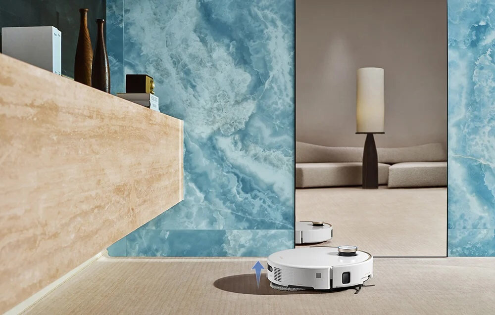 Biały robot sprzątający porusza się po jasnej podłodze w eleganckim wnętrzu z niebieską ścianą i dużym lustrem. W tle widać sofę i lampę stojącą na ciemnym stoliku.