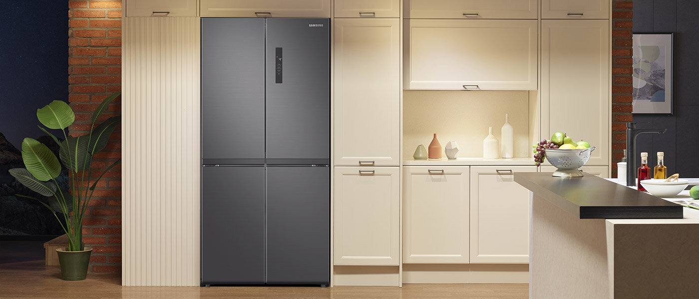Lodówka Samsung Multidoor z zewnętrznym wyświetlaczem doskonale komponuje się z nowoczesnym wystrojem kuchni, nadając jej elegancji i funkcjonalności. Grafitowa lodówka z dwoma drzwiami i wbudowanym dystrybutorem wody stoi w nowoczesnej kuchni. Obok znajdują się jasne szafki kuchenne i dekoracje, takie jak rośliny i miska z owocami.