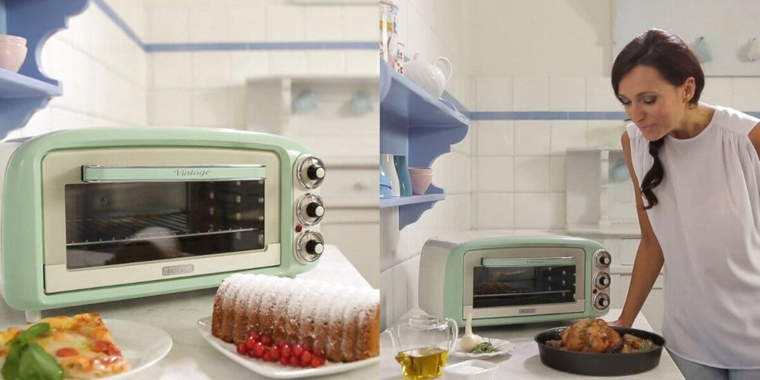 Na obrazie znajduje się mały piekarnik w stylu retro w kolorze miętowym z trzema pokrętłami. Obok piekarnika widoczne są potrawy, w tym ciasto i pieczone mięso, a w tle znajduje się osoba w białej bluzce.