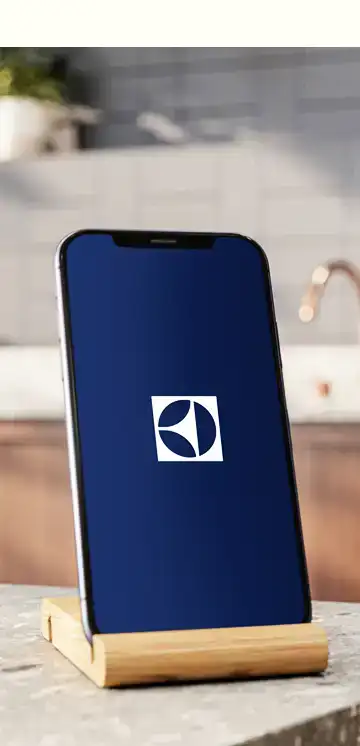 Smartfon na drewnianej podstawce z wyświetlonym logo Electrolux na ekranie. W tle widoczna jest kuchnia z szarymi kafelkami i kranem.