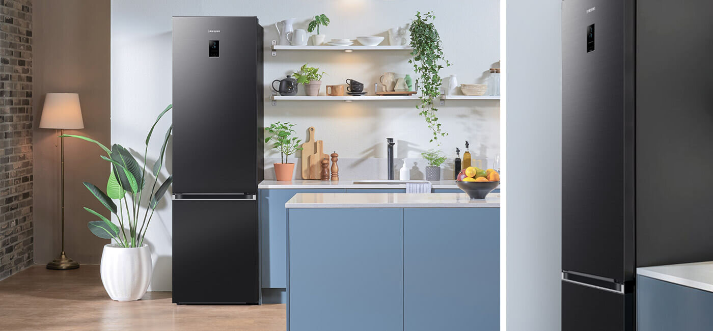 Design chłodziarko-zamrażarki Samsung RB38C774DB1 idealnie dostosowany do eleganckiego, nowoczesnego wnętrza W kuchni znajduje się grafitowa lodówka z wyświetlaczem na drzwiach. Obok lodówki są niebieskie szafki, a na blacie stoją rośliny i miska z owocami.