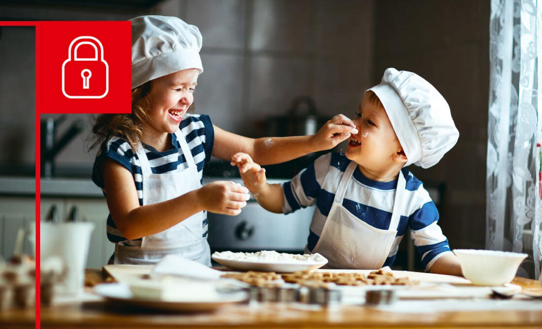 Dwoje dzieci w kucharskich czapkach i fartuchach bawi się przy stole pełnym składników do pieczenia. W lewym górnym rogu znajduje się czerwony symbol kłódki.