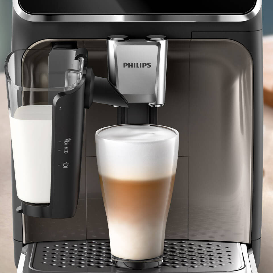 Ekspres do kawy z logo PHILIPS przygotowuje kawę z mleczną pianką w szklance. Obok znajduje się pojemnik na mleko z oznaczeniami poziomu napełnienia.