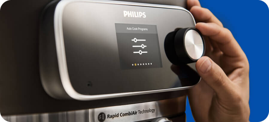 Osoba reguluje pokrętło na panelu sterowania urządzenia z wyświetlaczem pokazującym \'Auto Cook Programs\'. Widoczne jest logo \'PHILIPS\' oraz napis \'Rapid CombiAir Technology\'.