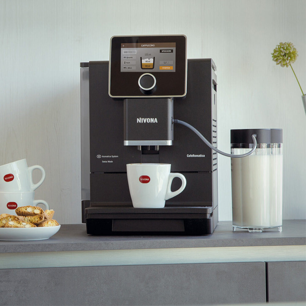 Na blacie stoi ekspres do kawy marki NIVONA z wyświetlaczem pokazującym opcje przygotowania cappuccino. Obok ekspresu znajduje się szklany pojemnik z mlekiem oraz filiżanki z logo NIVONA.