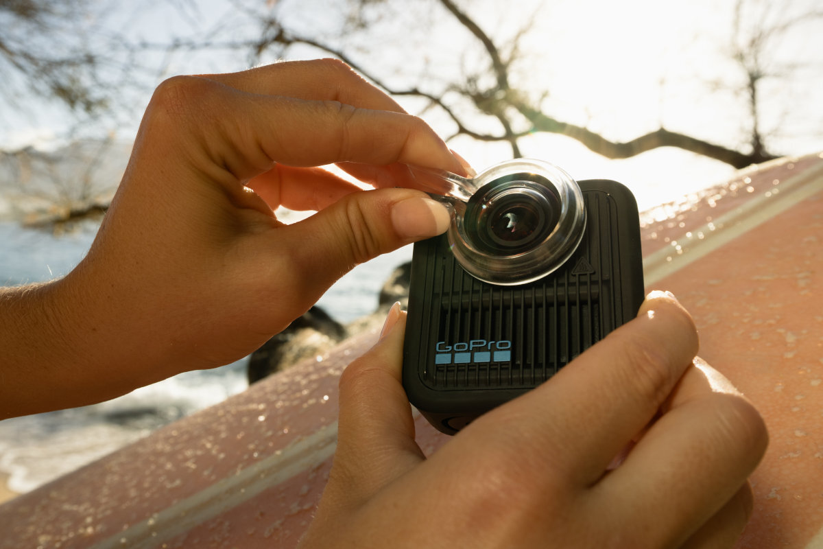 Osoba trzyma kamerę sportową z logo GoPro, zakładając osłonę na obiektyw. W tle widać rozmyte gałęzie drzewa i fragment deski surfingowej.